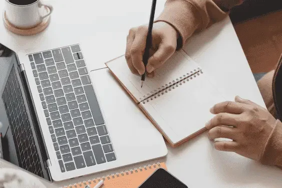 Image of a hand writing on a notepad in front of a laptop